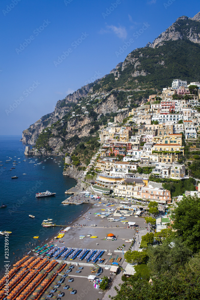 Positano, Amalfi Coast, Italy - high angle view shot on a sunny morning with blue skies, showing the houses on the hills, colorful umbrellas on the beach, and the boats near the shore