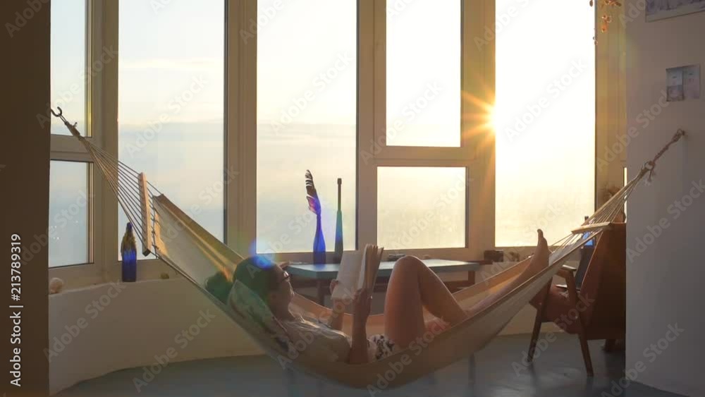 young female reading a book at home against panoramic window in hammok during sunrise or sunset young female reading a book at home against panoramic window in hammok during sunrise or sunset