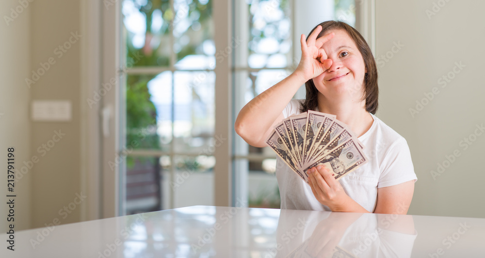 Down syndrome woman at home holding dollars with happy face smiling doing ok sign with hand on eye looking through fingers