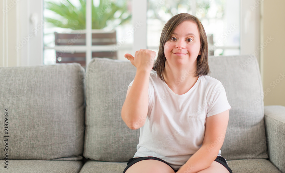 Down syndrome woman sitting on the sofa at home pointing and showing with thumb up to the side with happy face smiling