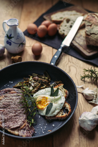 A view of a grilled beefsteak and an egg in a pan surrounded by eggs, garlic and olive oil