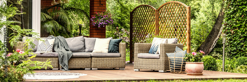 Panorama of wicker garden furniture with cozy pillows and blankets on a wooden terrace in beautiful outdoor greenery