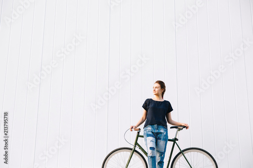 Fashionable beautiful girl standing with bicycle, looking away, over white wall background. Copy Space. Outdoors