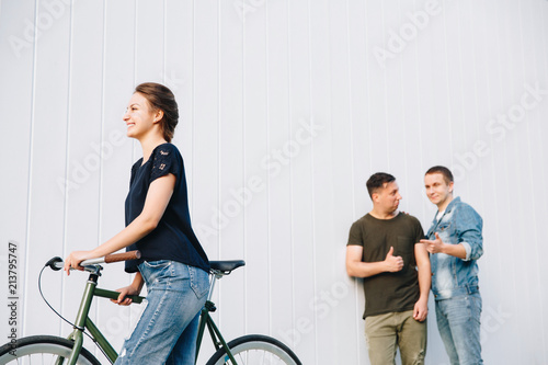 Two attractive young guys gossip about a woman, looking with enamored eyes at a single beautiful girl walking with bike, outdoors.