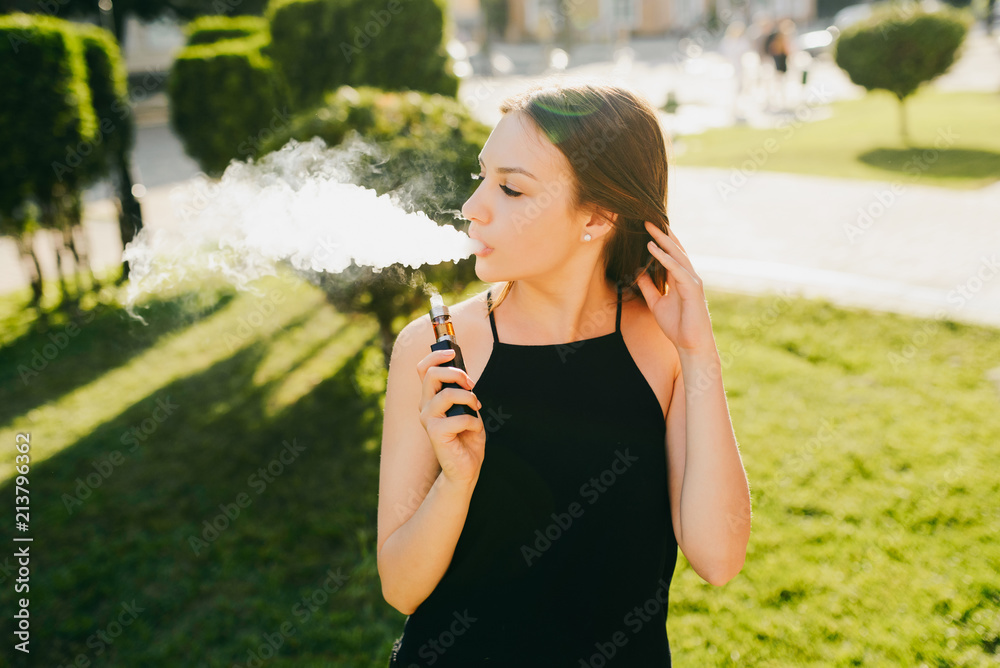Fototapeta premium Side view of young beautiful girl vaping e-cigarette, outdoors. Wearing black stylish singlet.
