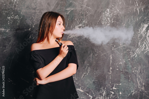 Fashionable young woman smoking the electronic cigarette. Dressed in black t-shirt standing over dark background. Indoors.