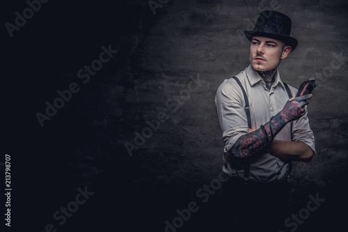 Portrait of a old-fashioned tattooed hairdresser wearing a white shirt with suspenders and cylinder hat, looking at a camera, holds a trimmer. Isolated on dark textured background.