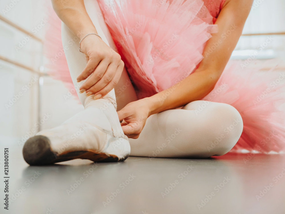 Young ballerina in pink tutu costume wraps white silk ribbons of soft top ballet shoes pointe and ties them up. Woman preparing for dance training lessons in gym.