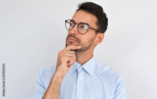 Closeup image of pleased pensive male keeps hand under chin, looking aside, on white studio background. Successful unshaven man thinks about creative project, wears blue shirt, copy space. People