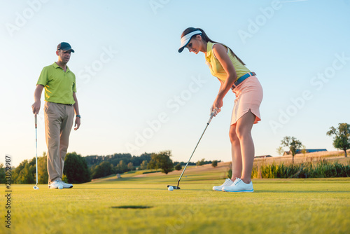 Full length of a woman calculating the trajectory of the ball to the hole, while playing professional golf with her male match partner or instructor outdoors in summer