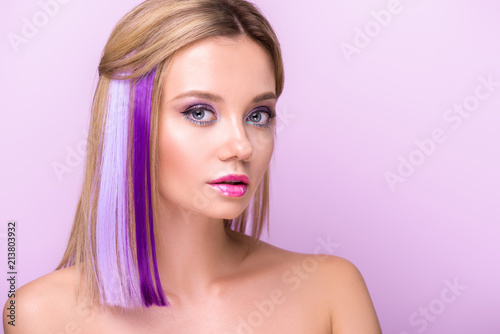 close-up portrait of stylish young woman with stylish makeup and purple hair strands looking at camera isolated on purple