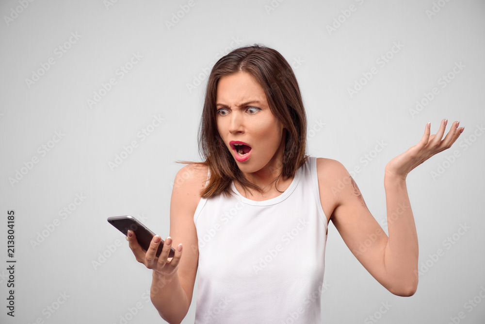 Studio shot of annoyed irritated angry woman. Worried with phone in hand feeling anxious and nervous. Portrait of a young brunette woman looking at the smartphone in her hand Studio shot of annoyed irritated angry woman. Worried with phone in hand feeling anxious and nervous. Portrait of a young brunette woman looking at the smartphone in her hand