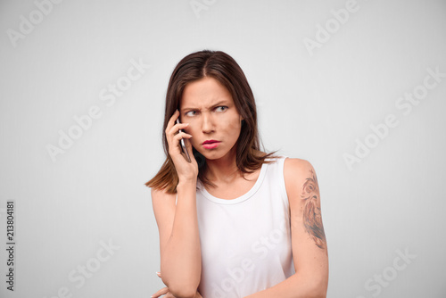 Indoor portrait of annoyed and angry woman with brunette hair looking aside while holding smartphone near ear, waiting when boyfriend will pick up phone, standing over gray background.