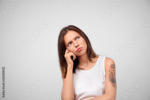 Horizontal portrait of attractive brunette female has unhappy look, thinks how to solve her problem, looks up, poses against white background with copy space