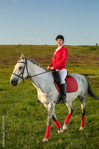 Young woman rider, wearing red redingote and white breeches, with her horse in evening sunset light.