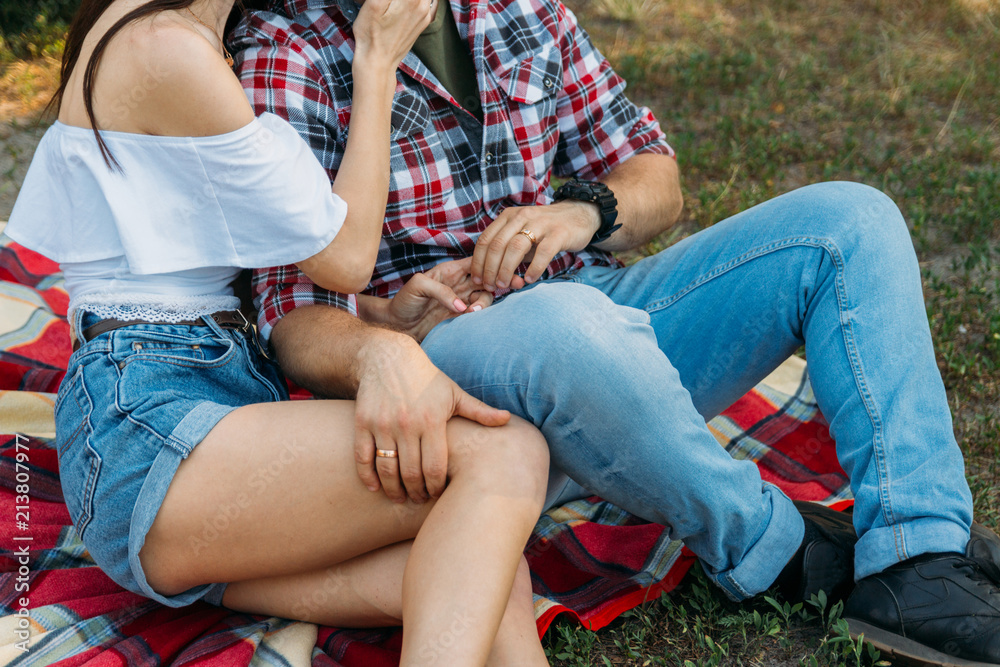 couple sitting on a plaid veil on the grass.the man put his hand on the girl's foot. a man in a plaid shirt and jeans, with a watch on his arm, a girl in a white blouse and denim shorts.
