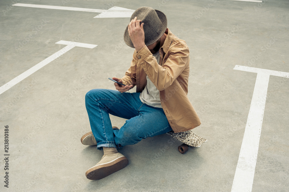 high angle view of stylish man in hat sitting on skateboard and using smartphone high angle view of stylish man in hat sitting on skateboard and using smartphone