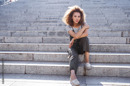 Photo of a girl with freckles and a haircut afro sitting on the staircase gazing and looking at the camera.