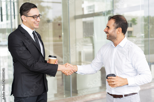 Portrait of happy male business colleagues shaking hands. Young manager in glasses greeting mid adult colleague at coffee break. Partnership concept