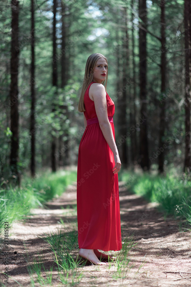 Blonde woman in red dress on forest path looking over shoulder.