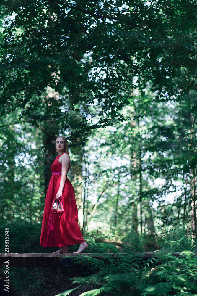 Woman in red dress crossing wooden bridge in summer forest.