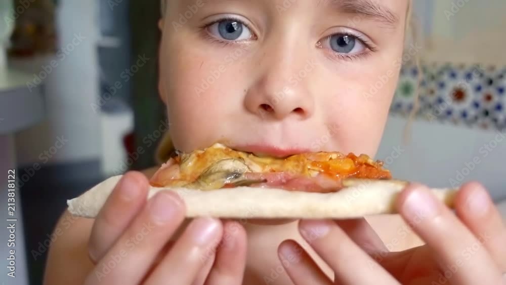 cute little girl is eating tasty pizza in home and looking at camera, close-up of her face cute little girl is eating tasty pizza in home and looking at camera, close-up of her face