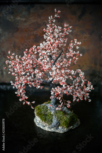 Bonsai tree of white and red beads and wire on a stone stand with moss on a metallic rust and black background. Homemade bonsai tree from beads