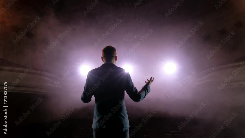 Showman. Young male entertainer, presenter or actor on stage. Back, arms to sides, smoke on background of spotlight. Rear view of a male public speaker speaking at the microphone, pointing, in the Showman. Young male entertainer, presenter or actor on stage. Back, arms to sides, smoke on background of spotlight. Rear view of a male public speaker speaking at the microphone, pointing, in the