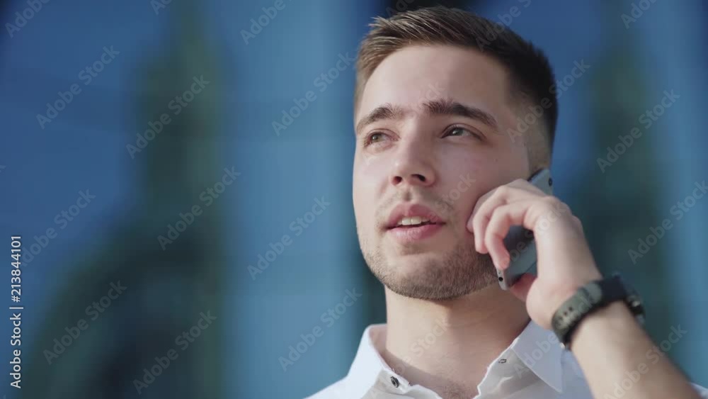 Pleasant young man in a white shirt speaks on the phone and smiles. He is located on the street, behind - a modern building. City Style. Portrait of a businessman with smartphone during speaking. Pleasant young man in a white shirt speaks on the phone and smiles. He is located on the street, behind - a modern building. City Style. Portrait of a businessman with smartphone during speaking.