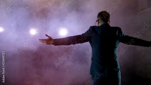 Showman. Young male entertainer, presenter or actor on stage. Back, arms to sides, smoke on background of spotlight. Rear view of a male public speaker speaking at the microphone, pointing, in the photo