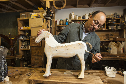 Business owner of antique restoration and wood carving studio at his workbench painting model of wooden horse in New York City, USA