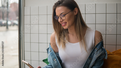 Young student girl surfing in internet using modern mobile phone during studying in cafeteria sitting near window photo