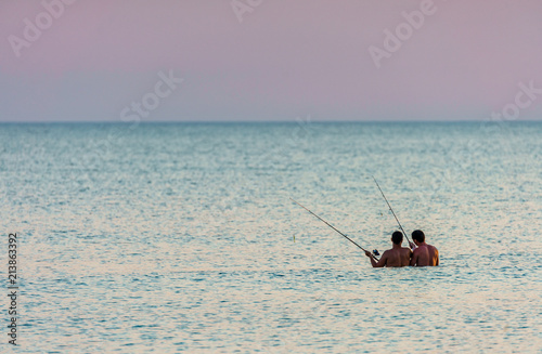 Couple of men holding fishing rods are fishing at sunset standing deep in sea water