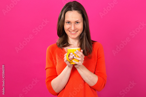 Happy brunette woman with a yellow cup in hands on a pink background. Drink hot drinks. Be healthy and not get sick.