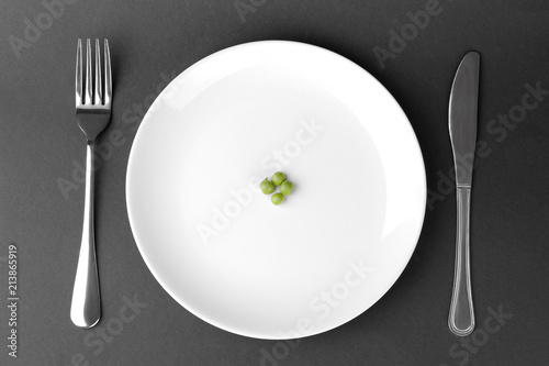 a few green fresh peas on a white plate next to a fork and knife on a black background. The concept of diet.