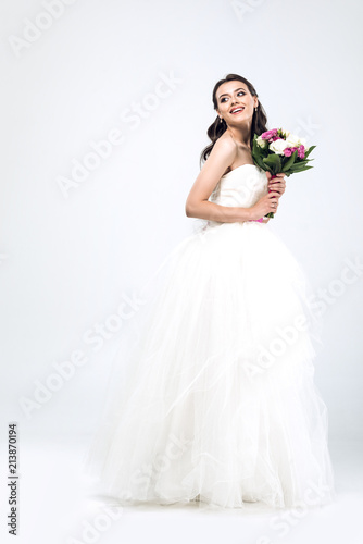 beautiful young bride in wedding dress with bouquet and looking away on white
