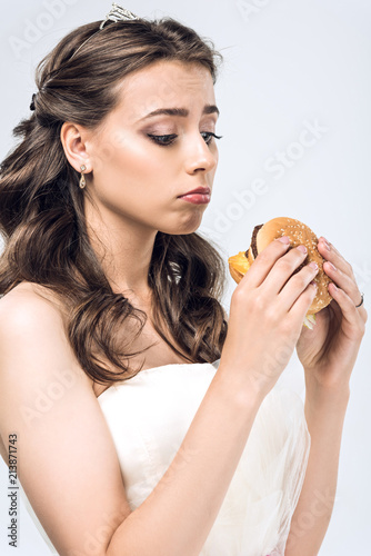 sad young bride in wedding dress looking at burger in hands isolated on white