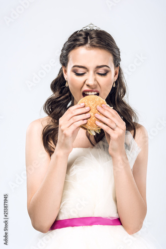 hungry young bride in wedding dress eating hamburger isolated on white