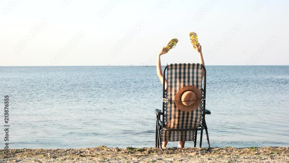 Rear view woman in beach chaise longue waving her flip-flops. Summer vacation and resort concept. Back view.