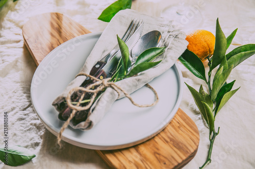 Table setting with white plate, cutlery, linen napkin and orange tree branch decoration on white linen tablecloth . Close up. Table with table setting and shair