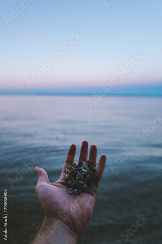 A man reaches his hand towards the water full of shells as the colors blend into the blue horizon.