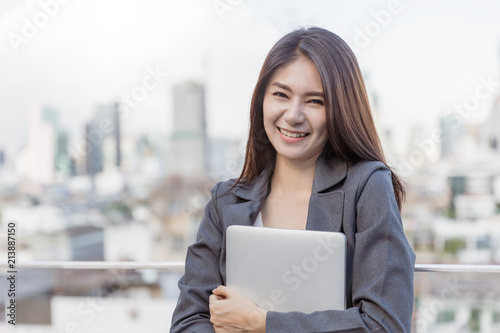 Business Woman Smile and holding computer laptop with office building background.