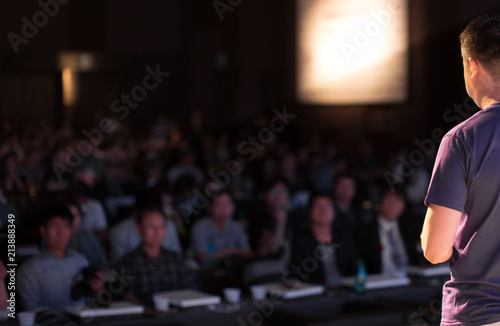 Presenter Speaking to Audience People in Conference Hall Auditorium. Presentation Stage. Blurred De-focused Unidentifiable Audience and Presenter. Technology. Casual Attire Presenter.