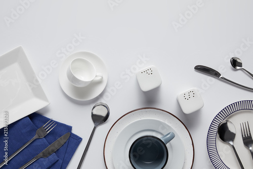top view of various ceramic plates, saltcellar, pepper caster, cup, kitchen towel, forks, spoons and knives on white table