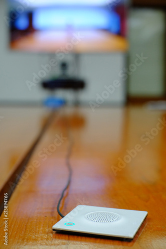 Microphone for teleconference system set standing on wood table with monitor on background. Selective focus.