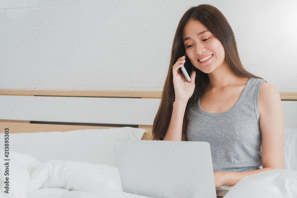 Asian beautiful smiling women using laptop while talking with smartphone on white bed in bedroom interior morning.Concept of people use technology. Asian beautiful smiling women using laptop while talking with smartphone on white bed in bedroom interior morning.Concept of people use technology.