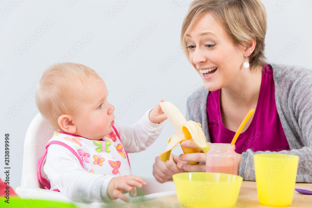 Portrait of a happy mother giving a fresh and nutritious banana to her cute 6 month old baby girl, while starting to eat solid food at home Portrait of a happy mother giving a fresh and nutritious banana to her cute 6 month old baby girl, while starting to eat solid food at home