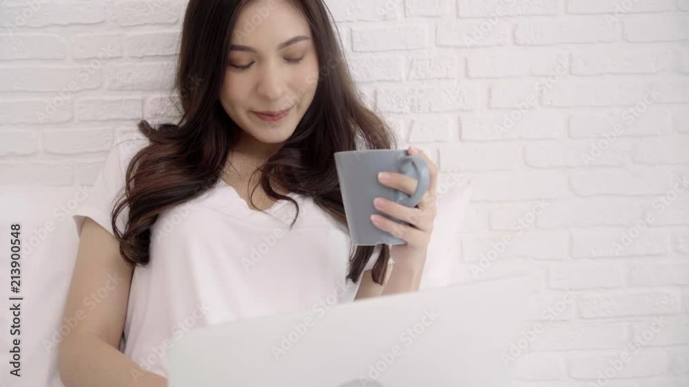 Beautiful Asian woman playing computer or laptop while lying on the bed in her bedroom. Happy female drinking coffee while buying online at home. Lifestyle woman at home concept. Beautiful Asian woman playing computer or laptop while lying on the bed in her bedroom. Happy female drinking coffee while buying online at home. Lifestyle woman at home concept.