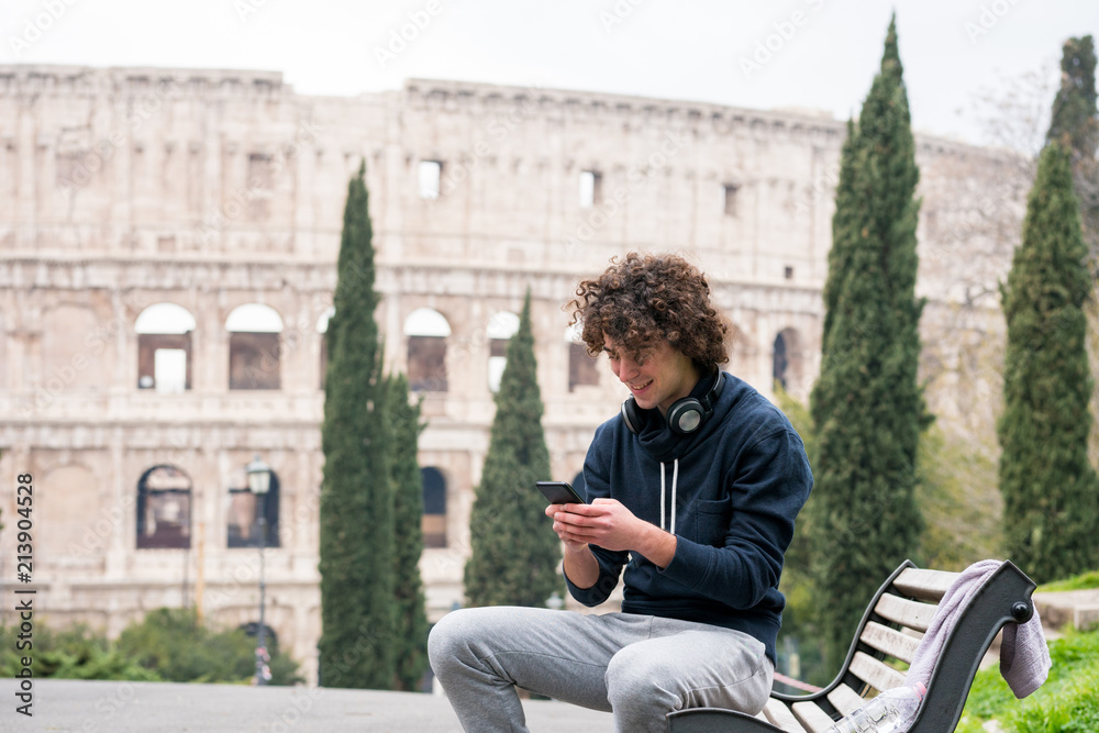 Handsome young man in sports cloths using his mobile phone while sitting on a bench in Rome. Young sportsman in tracksuit relaxing after training with Colosseum in the background Handsome young man in sports cloths using his mobile phone while sitting on a bench in Rome. Young sportsman in tracksuit relaxing after training with Colosseum in the background