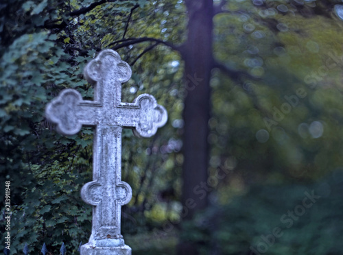 Obraz na plátně Stone grave cross on the blurred background of a tree leaves at midnight time wi